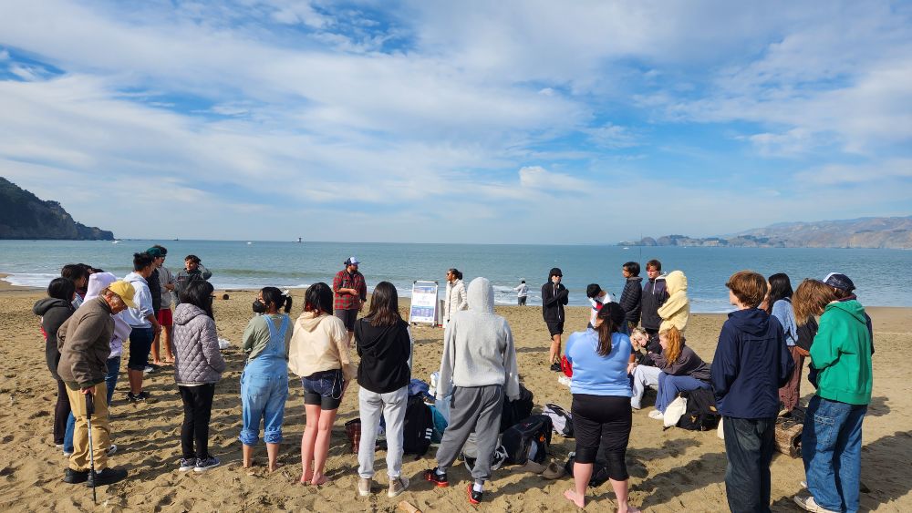 students gather around a sanctuary intern teaching on a sandy beach with the ocean behind them